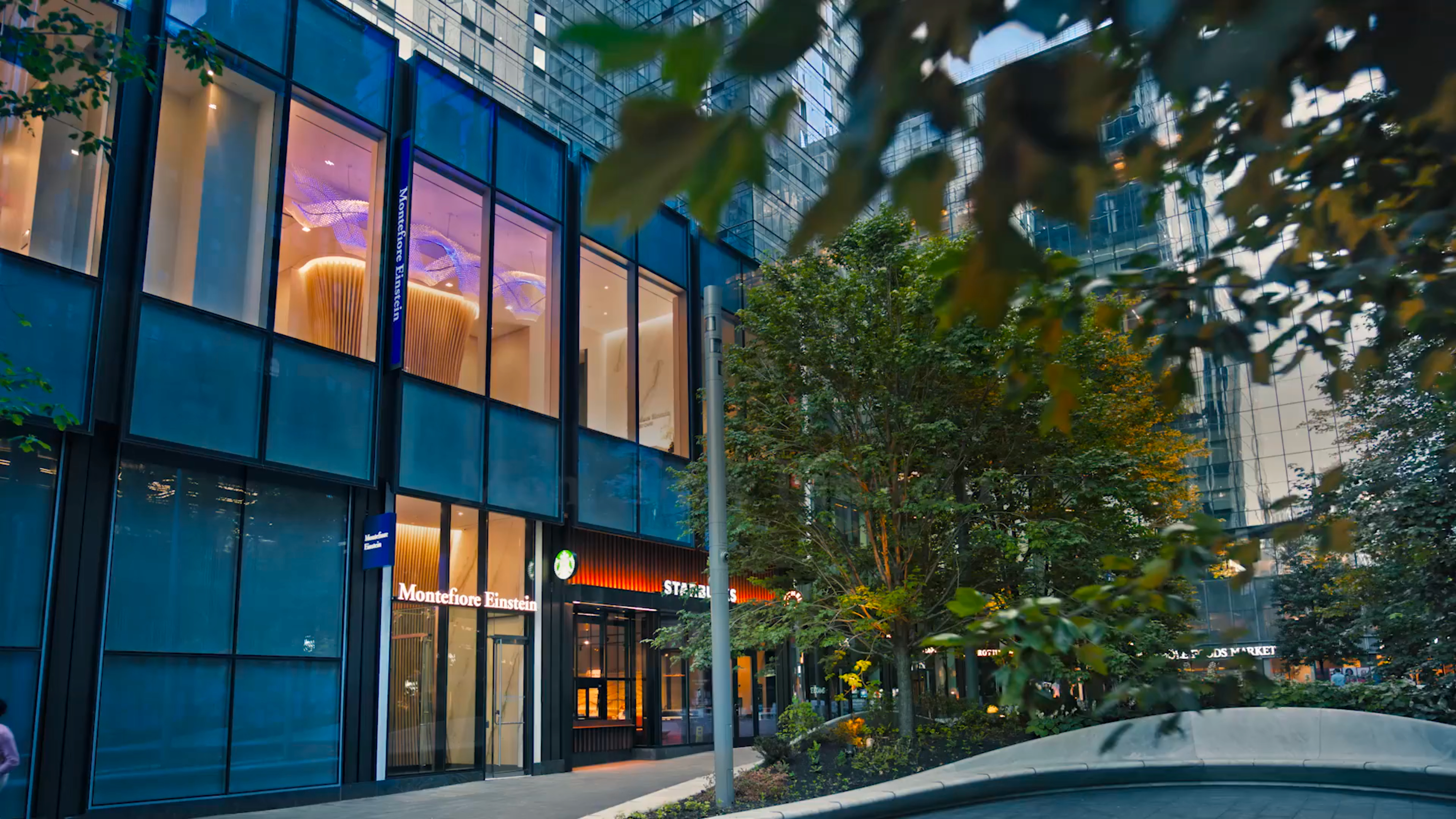 Exterior of Montefiore Einstein Manhattan West, a modern glass-facade building with illuminated interior, located at street level in a mixed-use development.