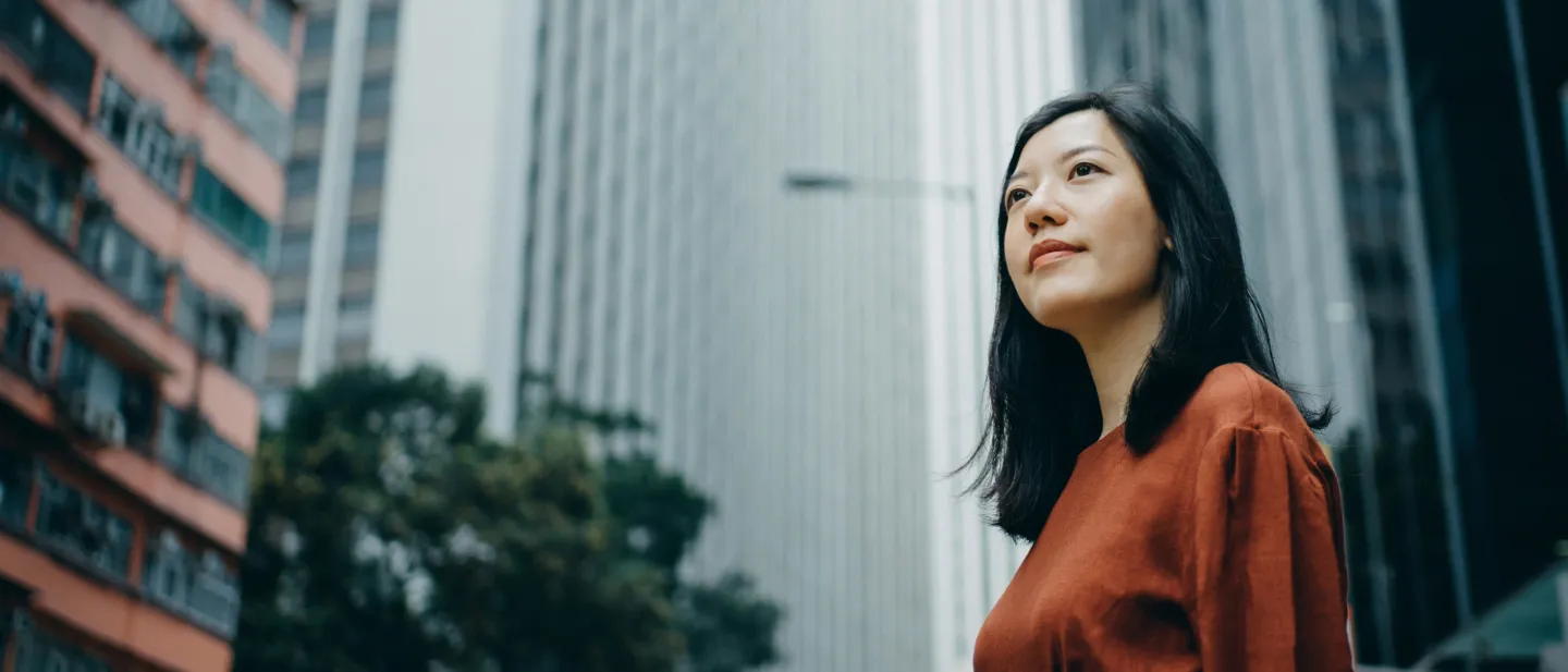 Woman amid tall buildings looking upwards.