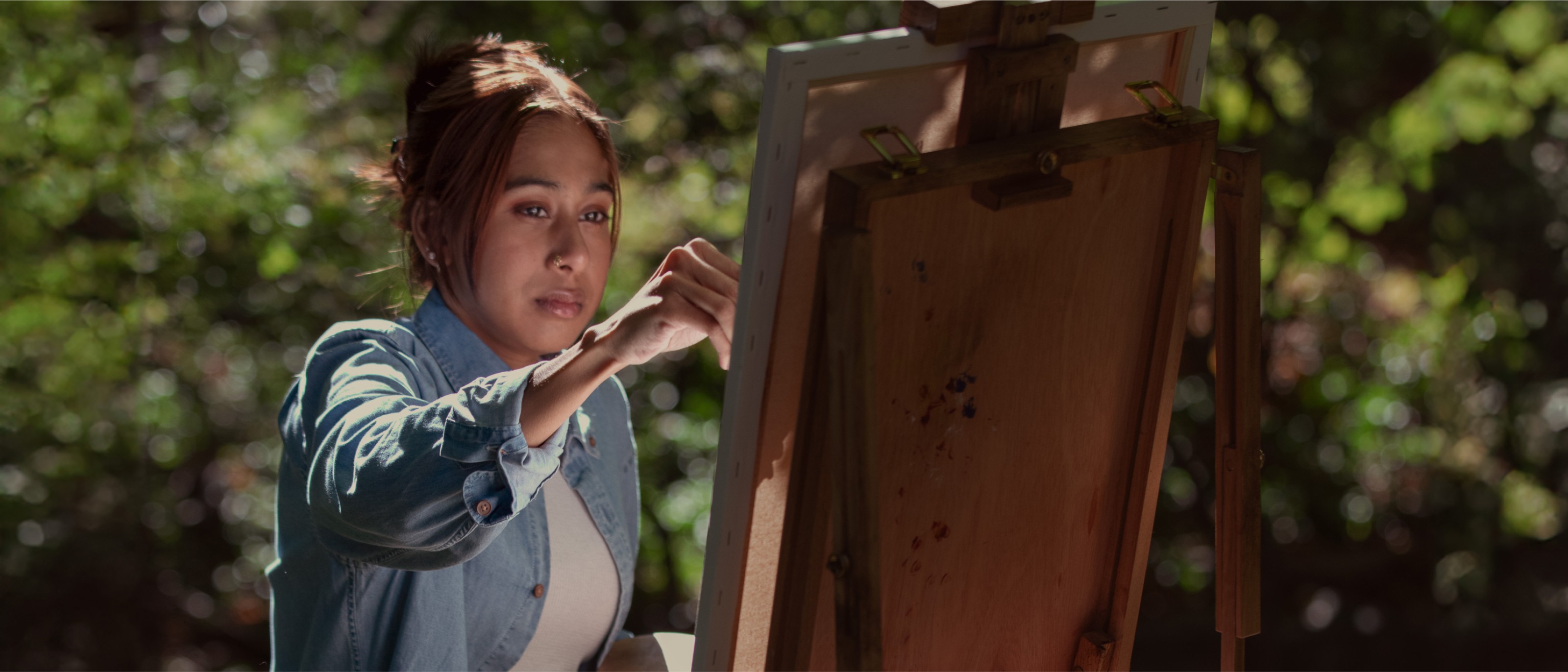 Alisha Khan seated outdoors on a bright day painting a canvas on an easel.