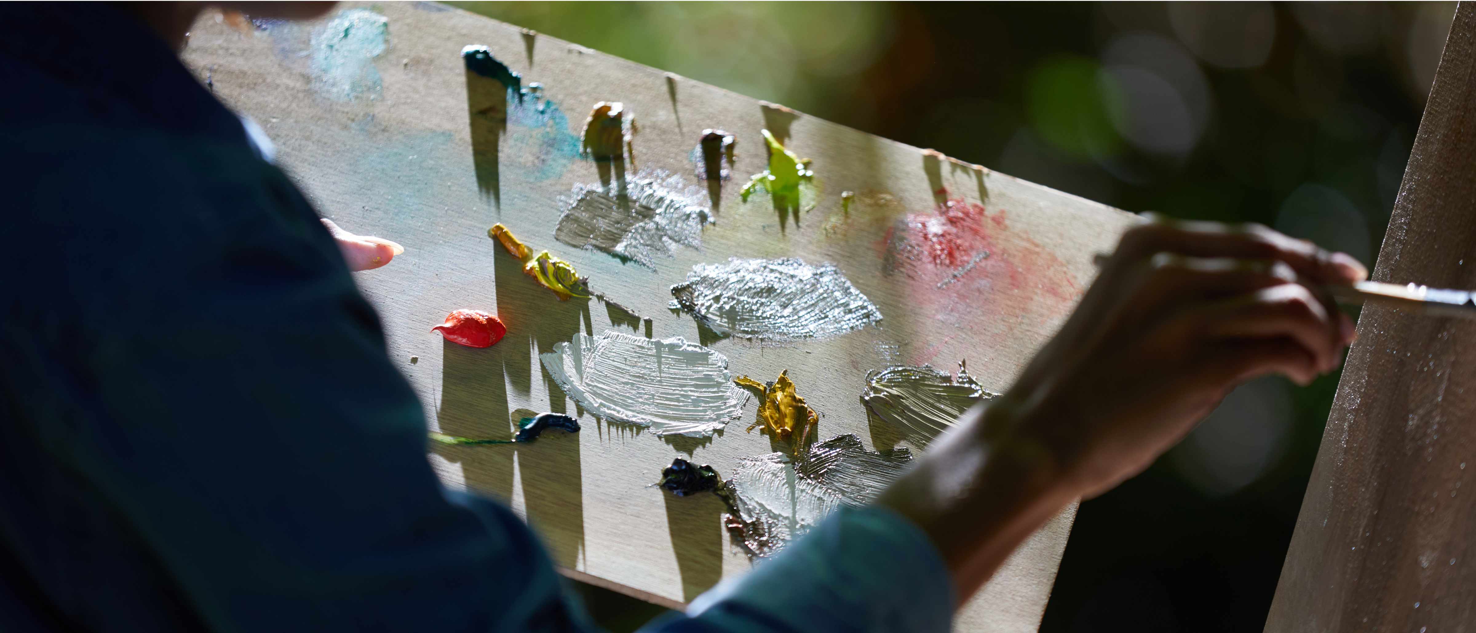 Close-up of Alisha Khan painting canvas while holding wood palette full with colored paint.