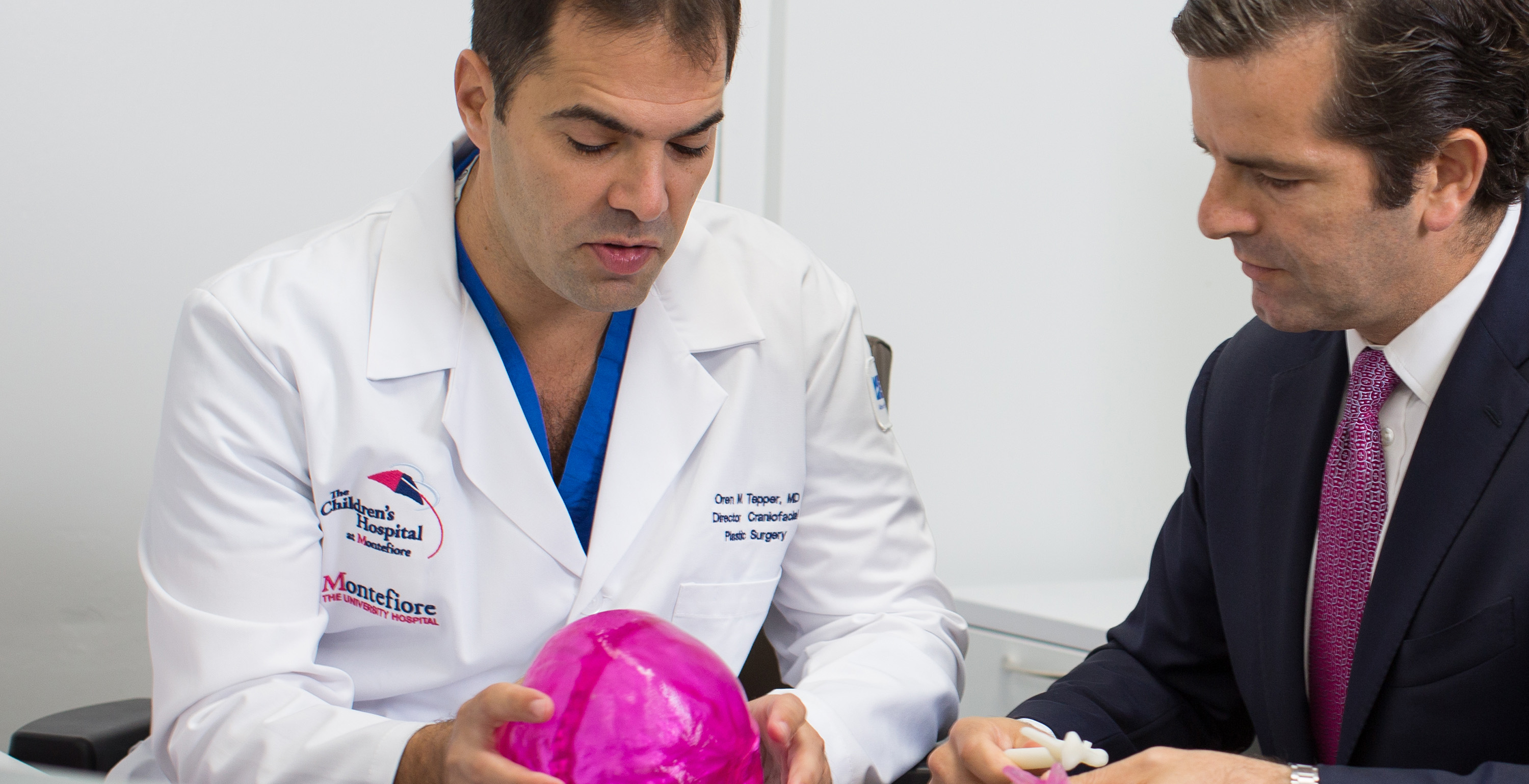 Doctor in white coat discussing a bright pink anatomical model with a suited man in a medical office.