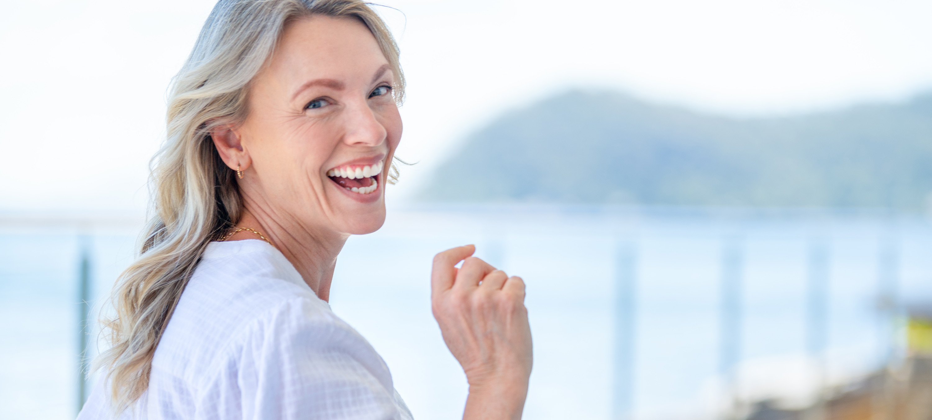 Joyful woman with light hair smiling at the camera while standing near a scenic waterfront.