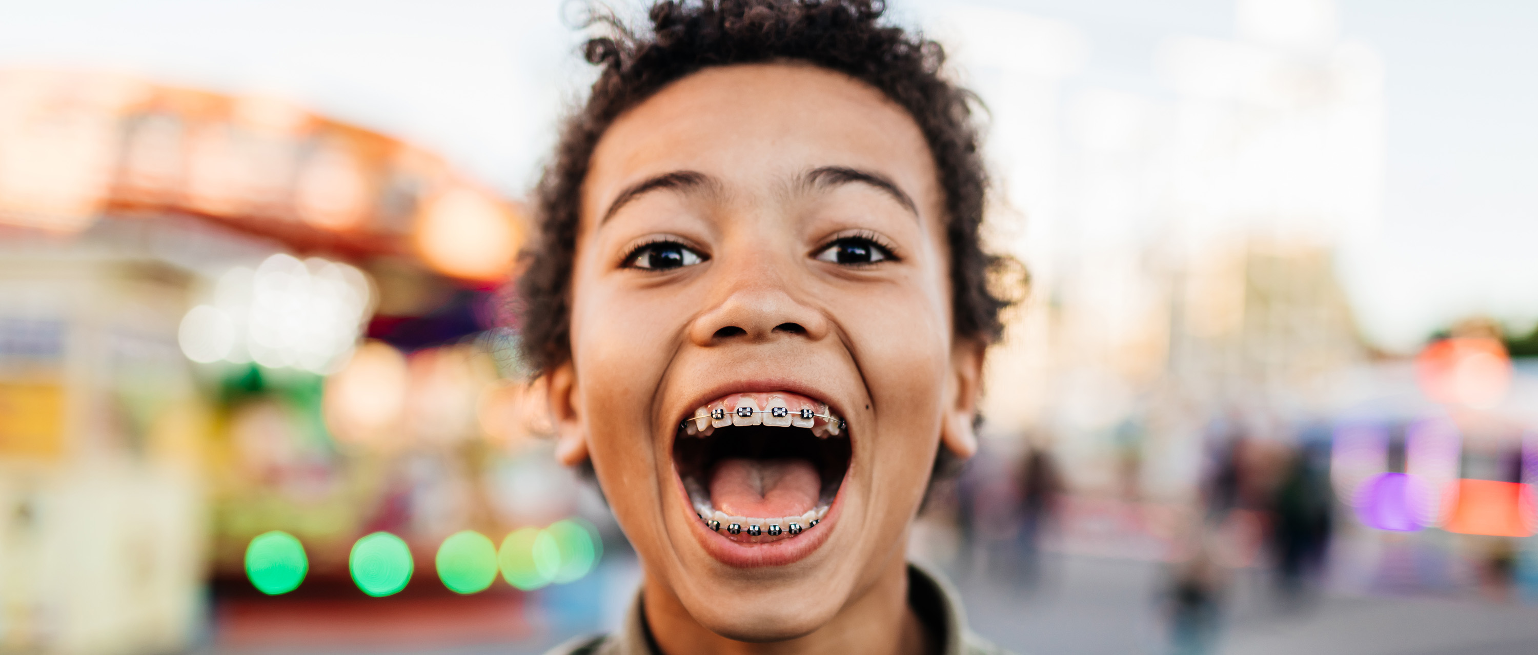 Excited boy smiling wide with braces at a colorful amusement park.