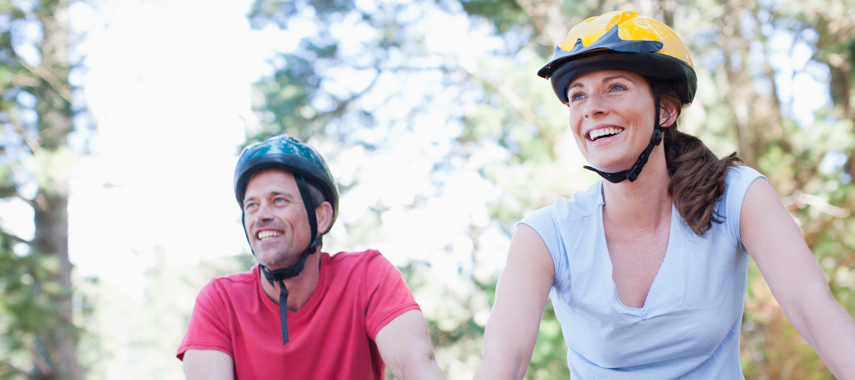 Couple wearing bike helmets smiling while riding through a sunlit forest.