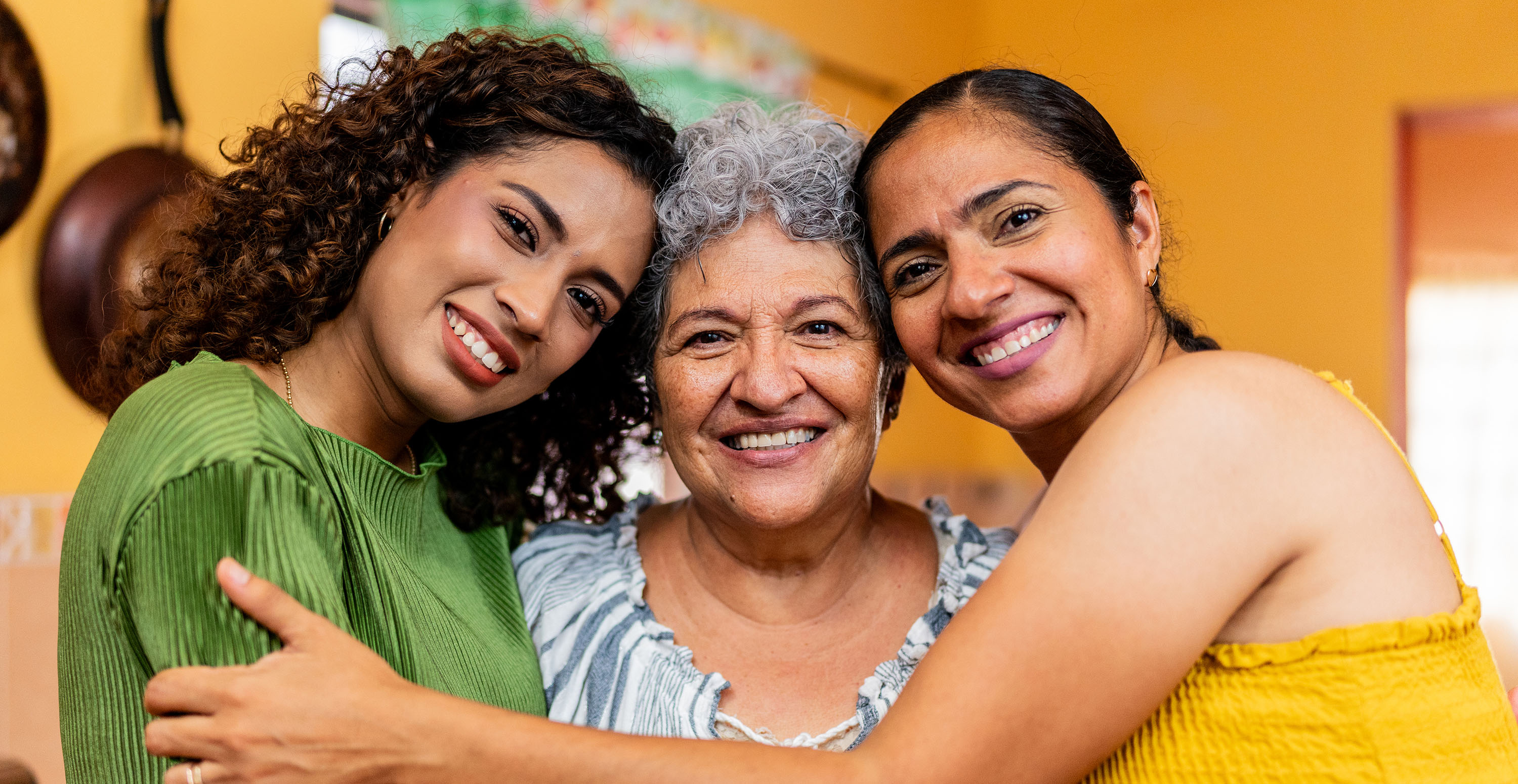 Three generations of women hug and smile together in a warmly lit kitchen.