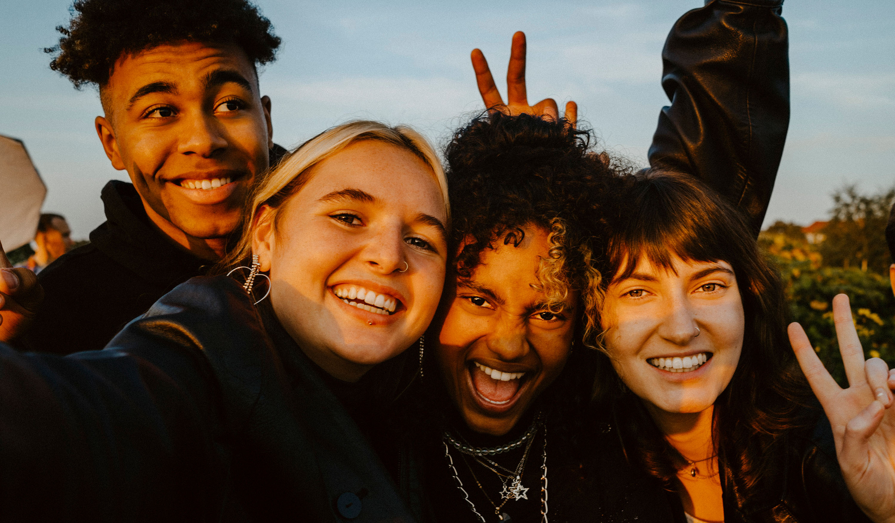Group of four friends smiling and taking a selfie outdoors in golden light.