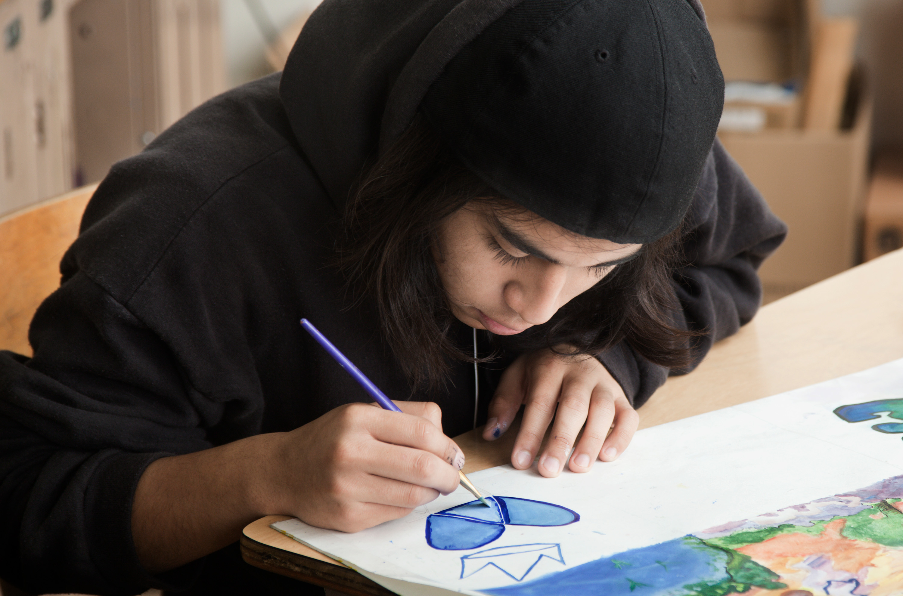 Teen in hoodie focused on painting a blue flower at a desk in classroom.