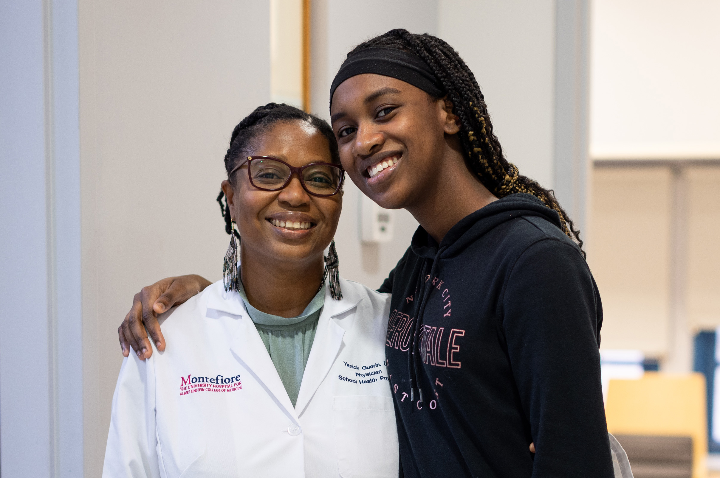 Teen smiles with her arm around a doctor in a white coat inside a clinic or hospital.