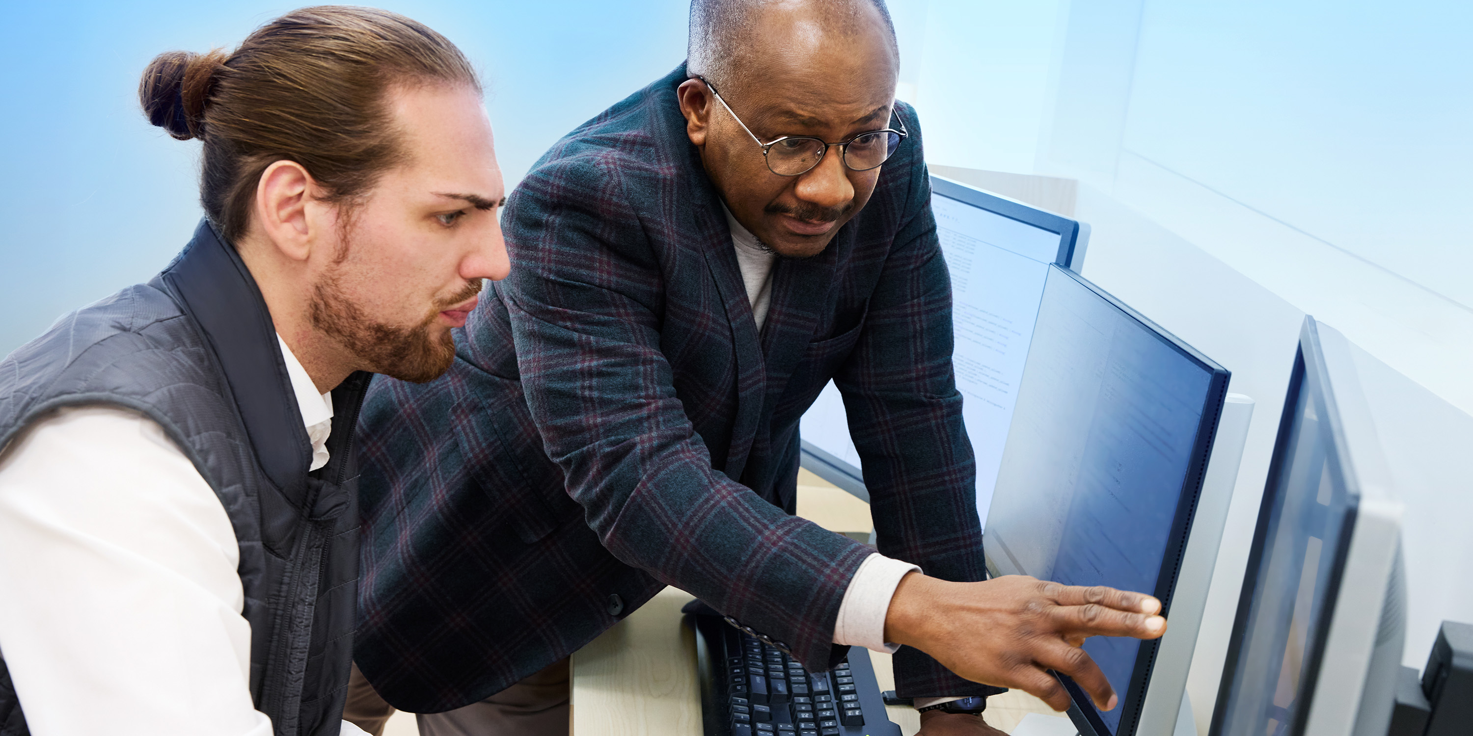 Two men collaborate at a computer workstation, with one pointing at the screen while explaining something to the other.