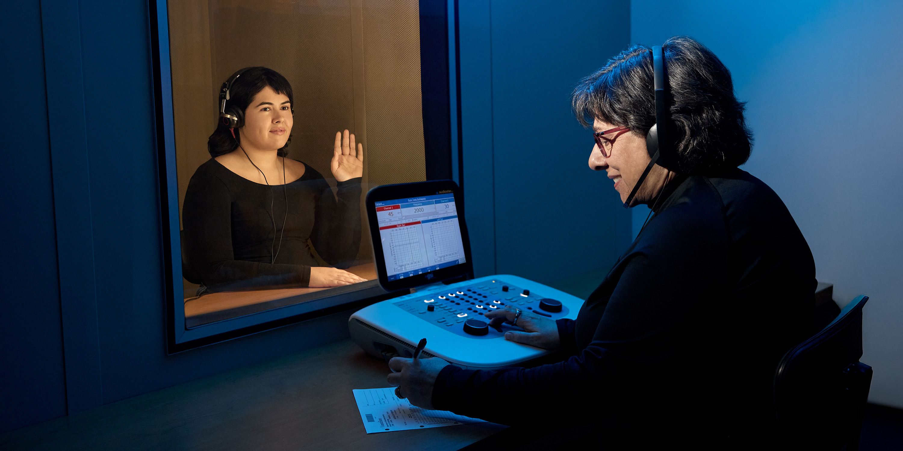 Audiologist conducting a hearing test with a patient seated behind glass, both wearing headsets.