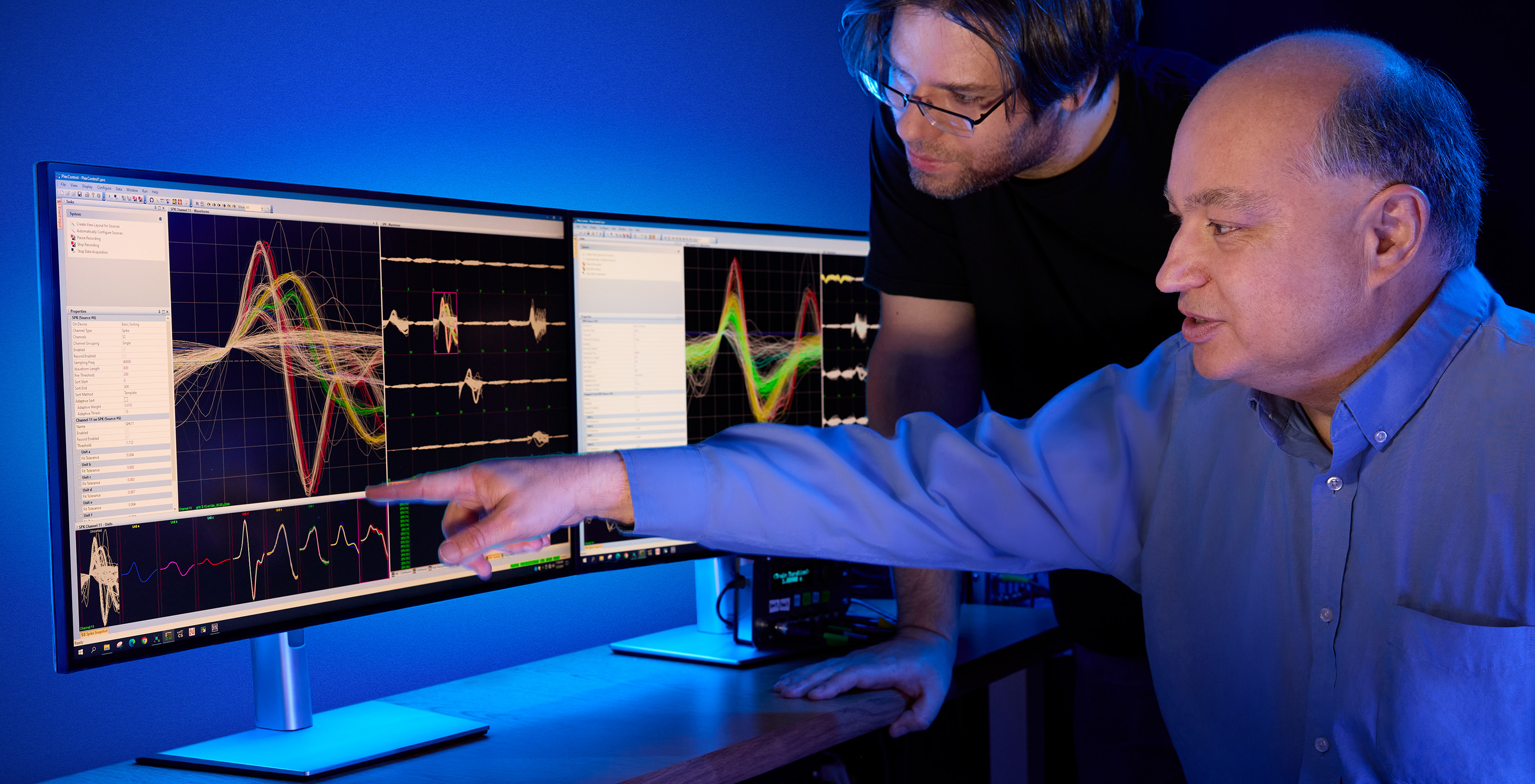 Two researchers analyzing colorful waveforms on dual monitors in a dimly lit lab with a blue light glow.
