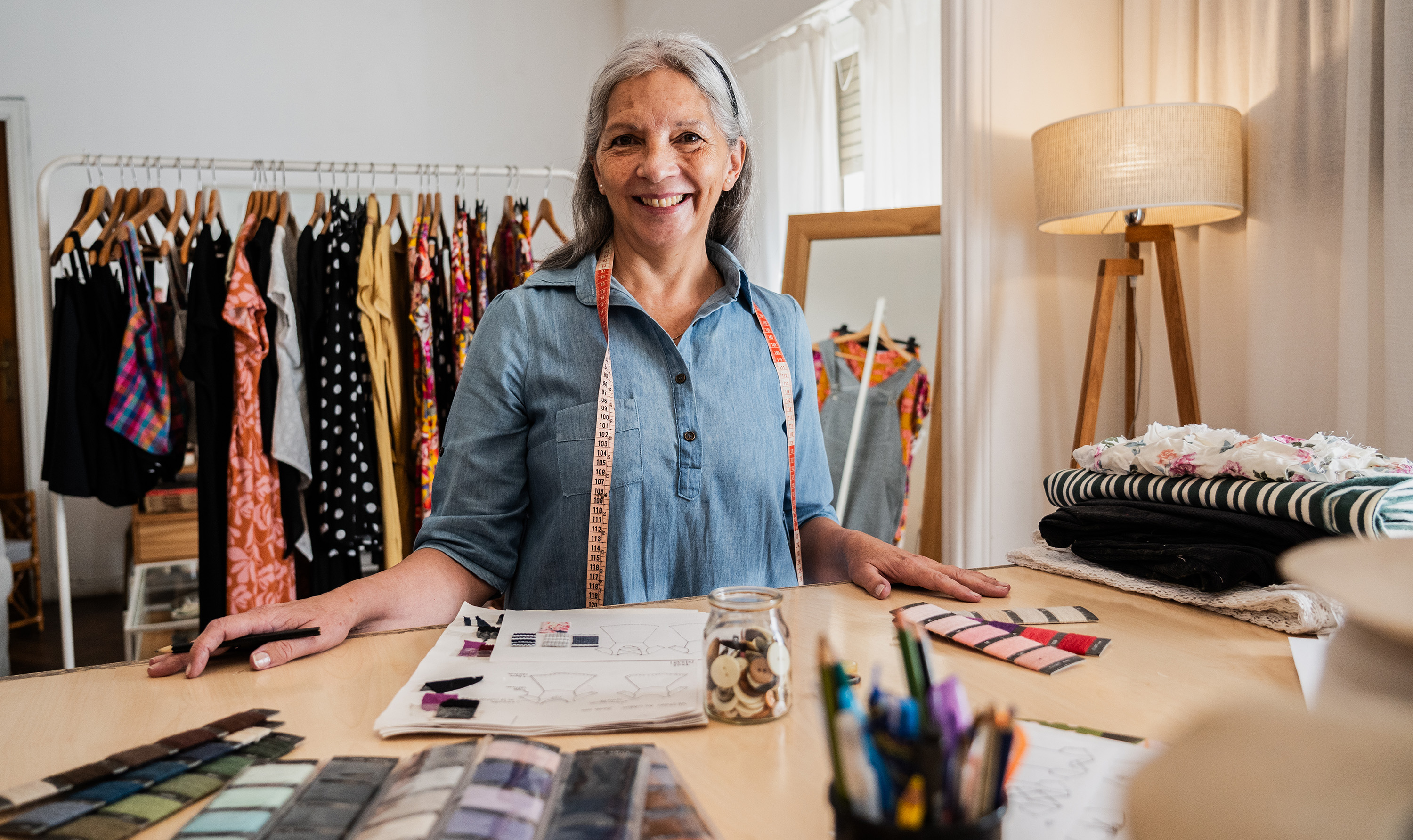 Smiling woman standing behind a table with sewing supplies in a clothing studio, surrounded by colorful garments and fabric swatches.