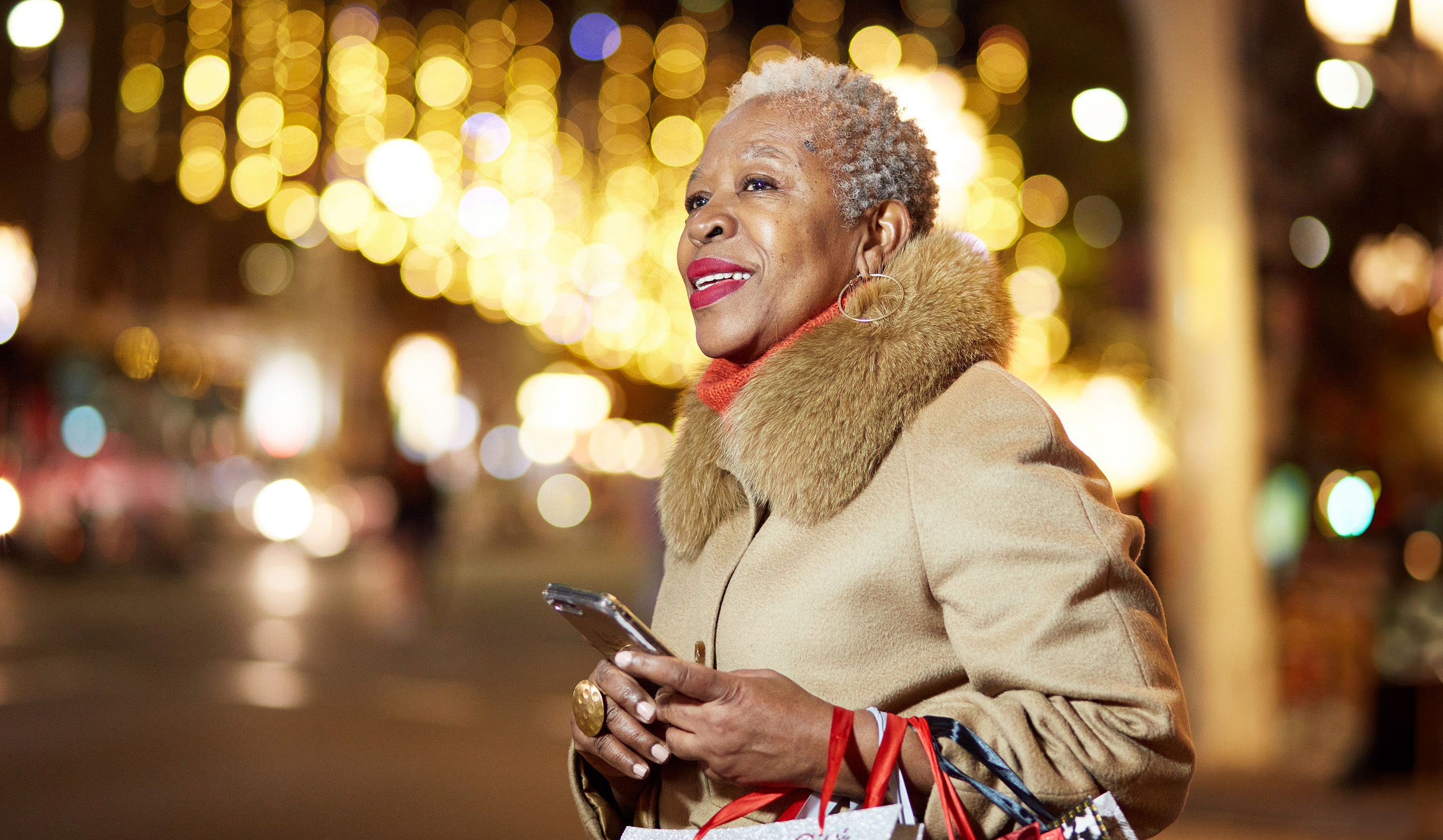 Stylish older woman holding a smartphone and shopping bags, smiling while standing on a city street at night with festive lights in the background.