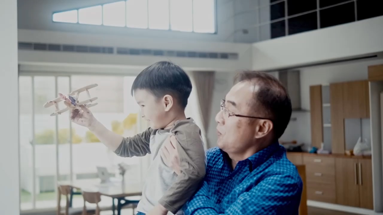 Grandfather holding young boy playing with a wooden toy airplane in a sunlit modern living room.