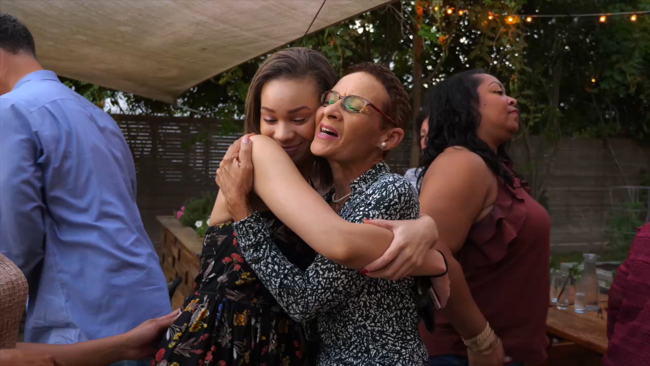 Two women share an emotional embrace at an outdoor gathering, surrounded by friends and string lights in a backyard setting.