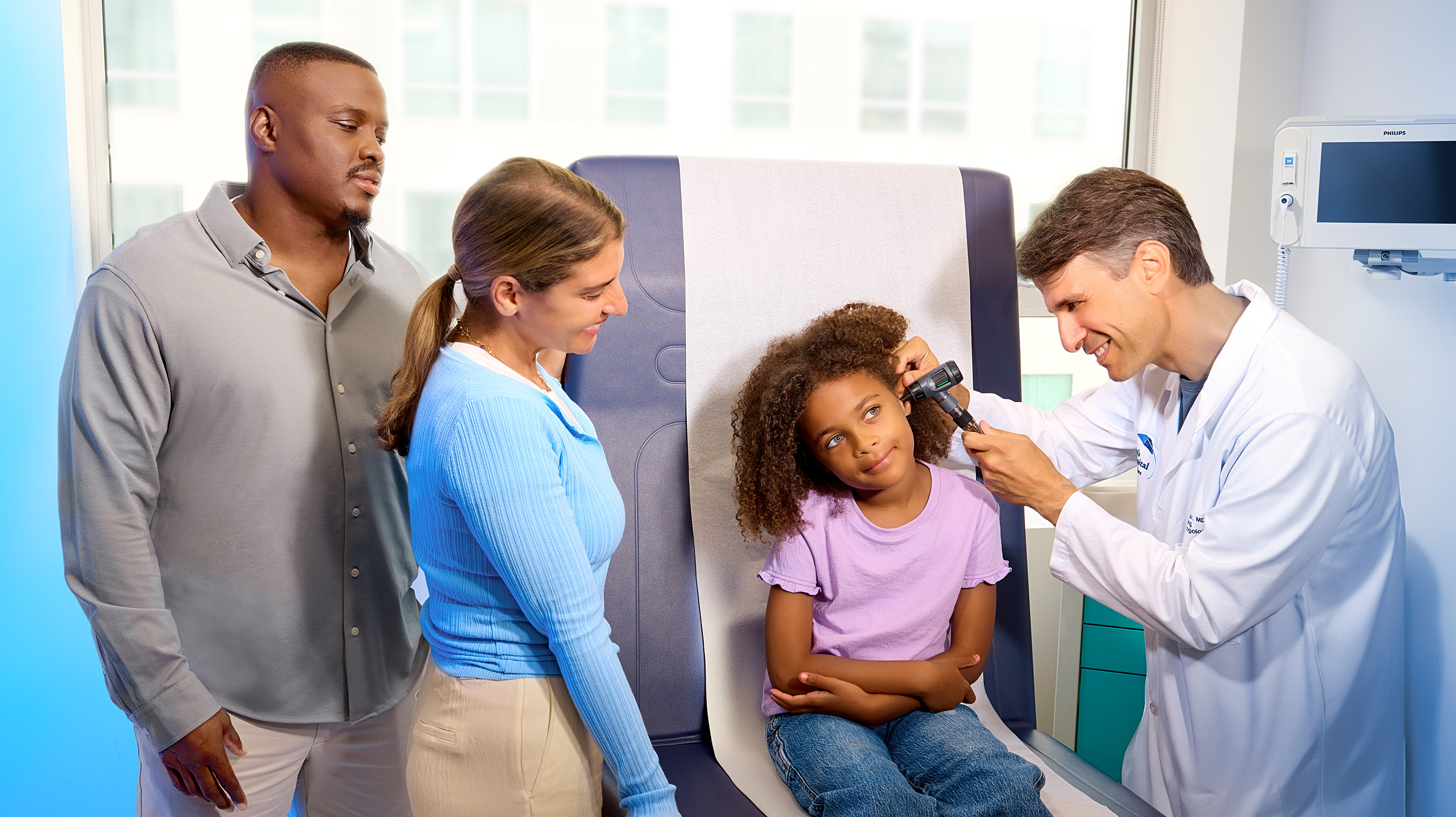 Pediatrician examining a young girl’s ear with an otoscope while her parents stand nearby, smiling and engaged in a bright clinic exam room.