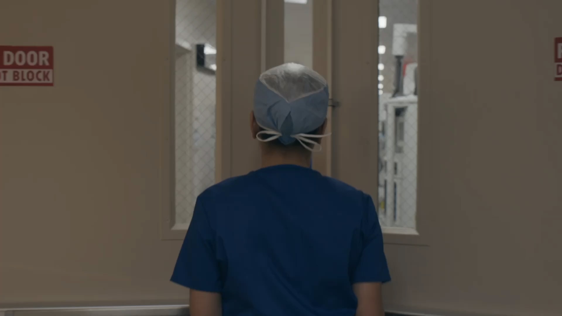 Healthcare worker in blue scrubs stands facing closed double doors in a hospital corridor