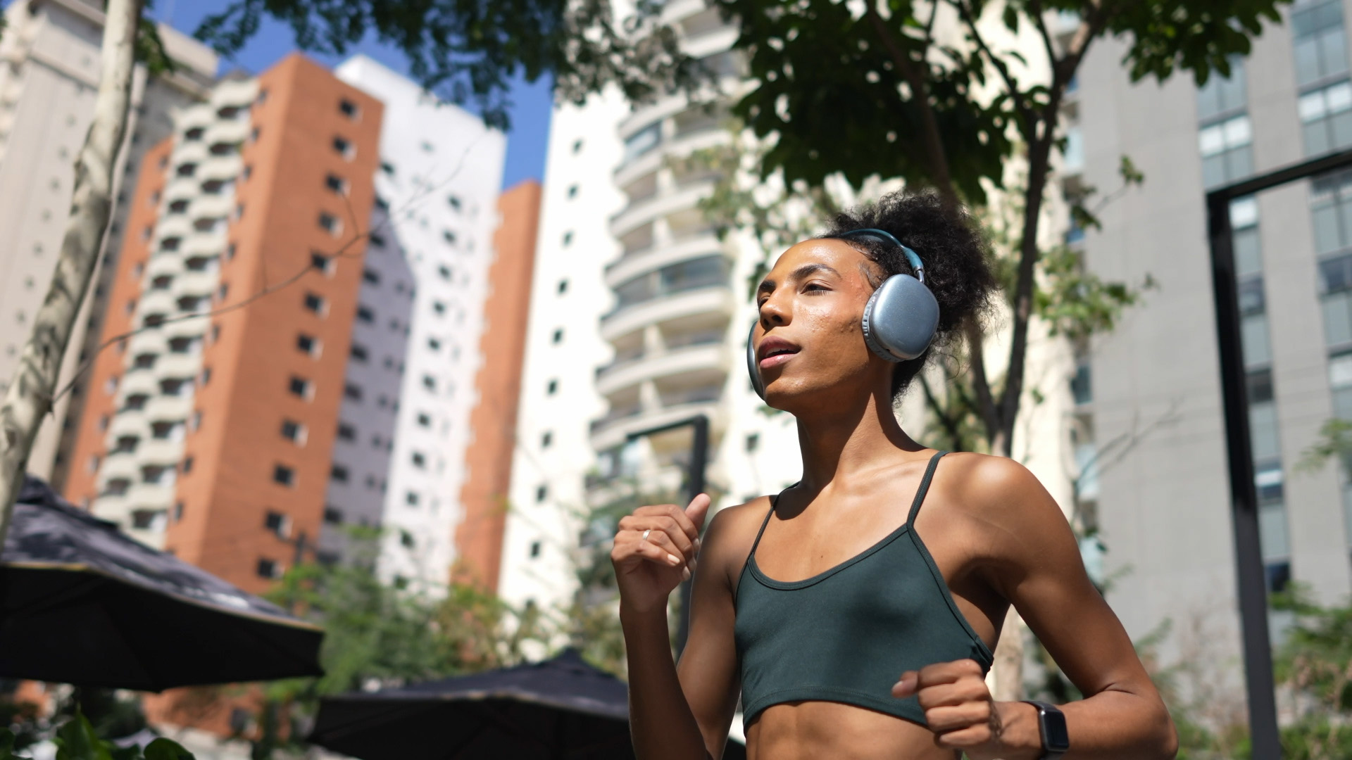 Trans woman jogging outdoors with headphones, wearing a sports bra, with tall city buildings in the background.