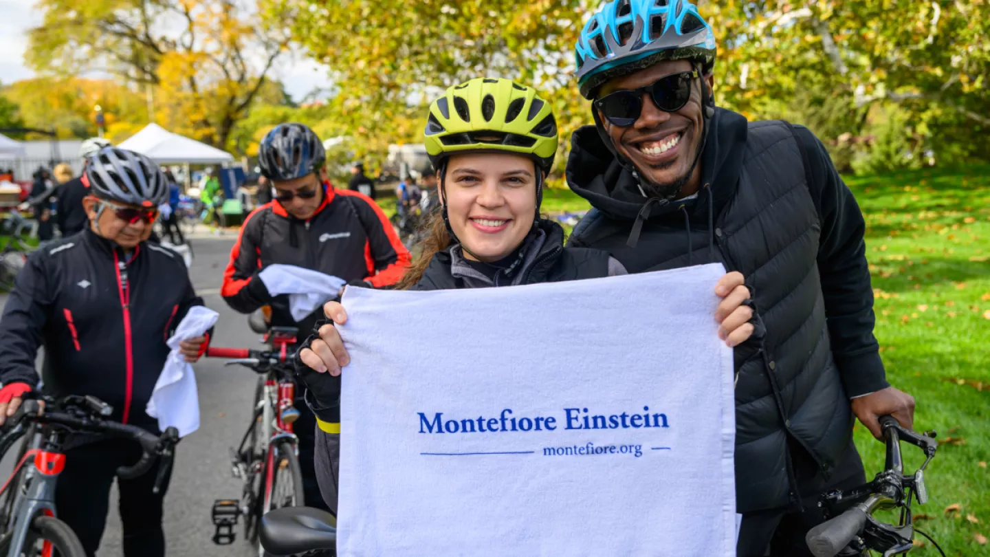 Two smiling bicyclists getting ready for race holding up a Montefiore Einstein towel