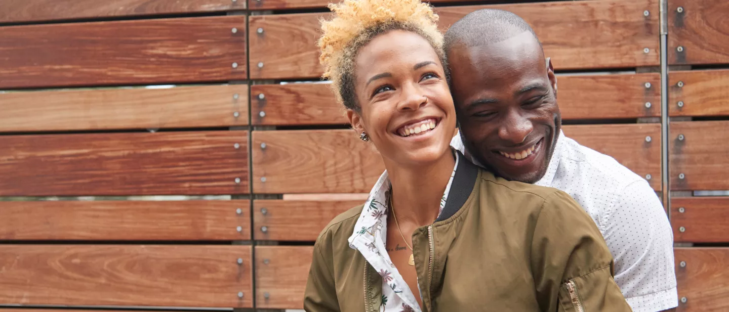 A smiling black couple embraces in front of a wall of horizontal wood slats of varying widths.