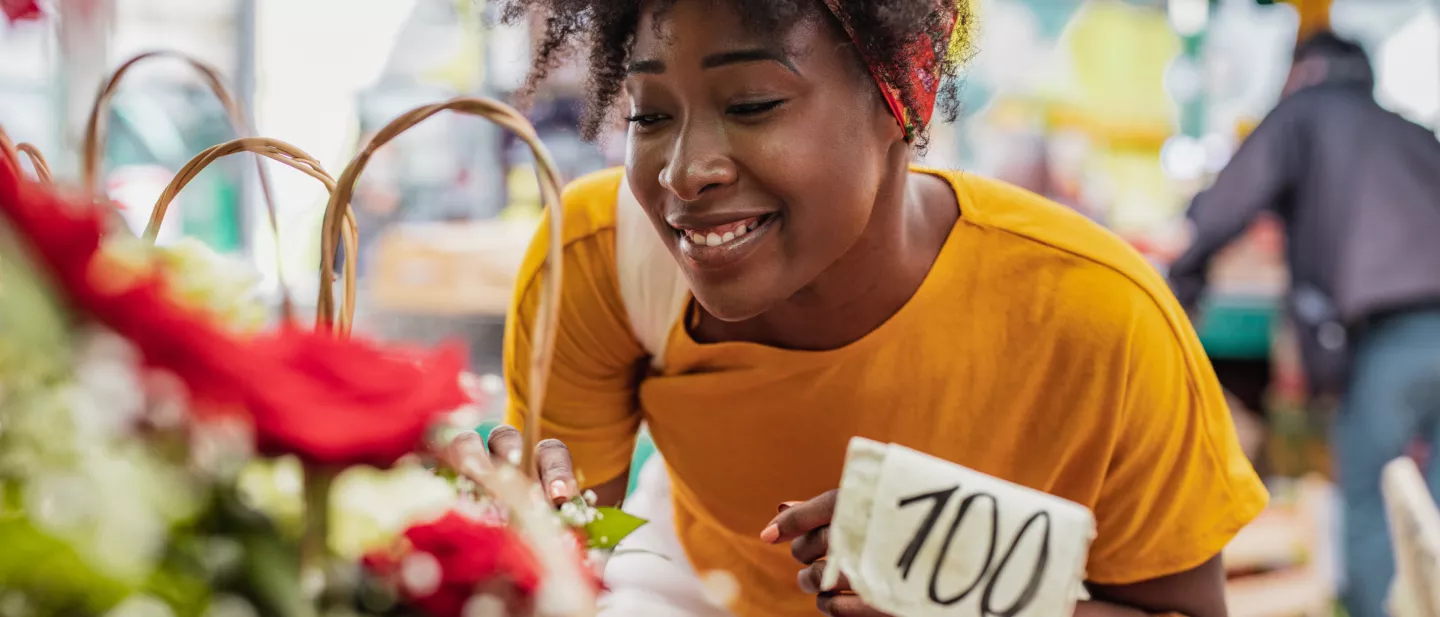 A happy black woman in a bright yellow top and red bandana browses flowers in an open-air market.