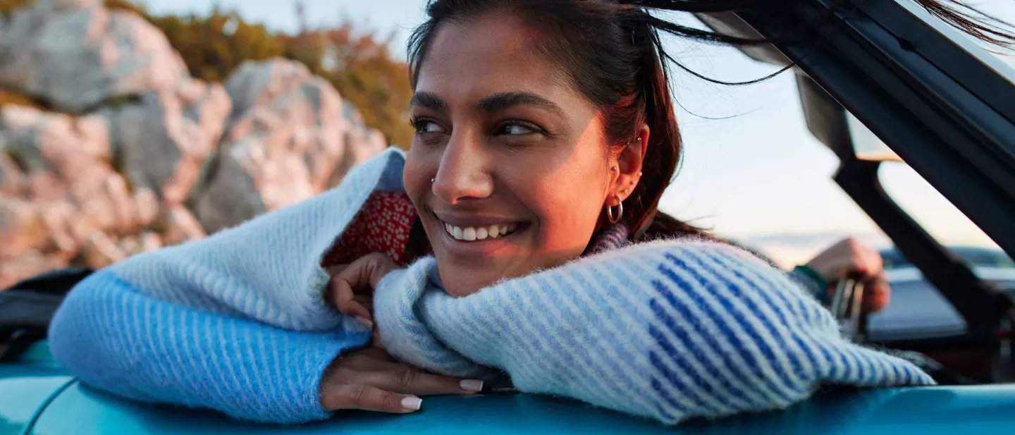Smiling female in convertible car parked at beach with arms folded in car leaning out passenger side window
