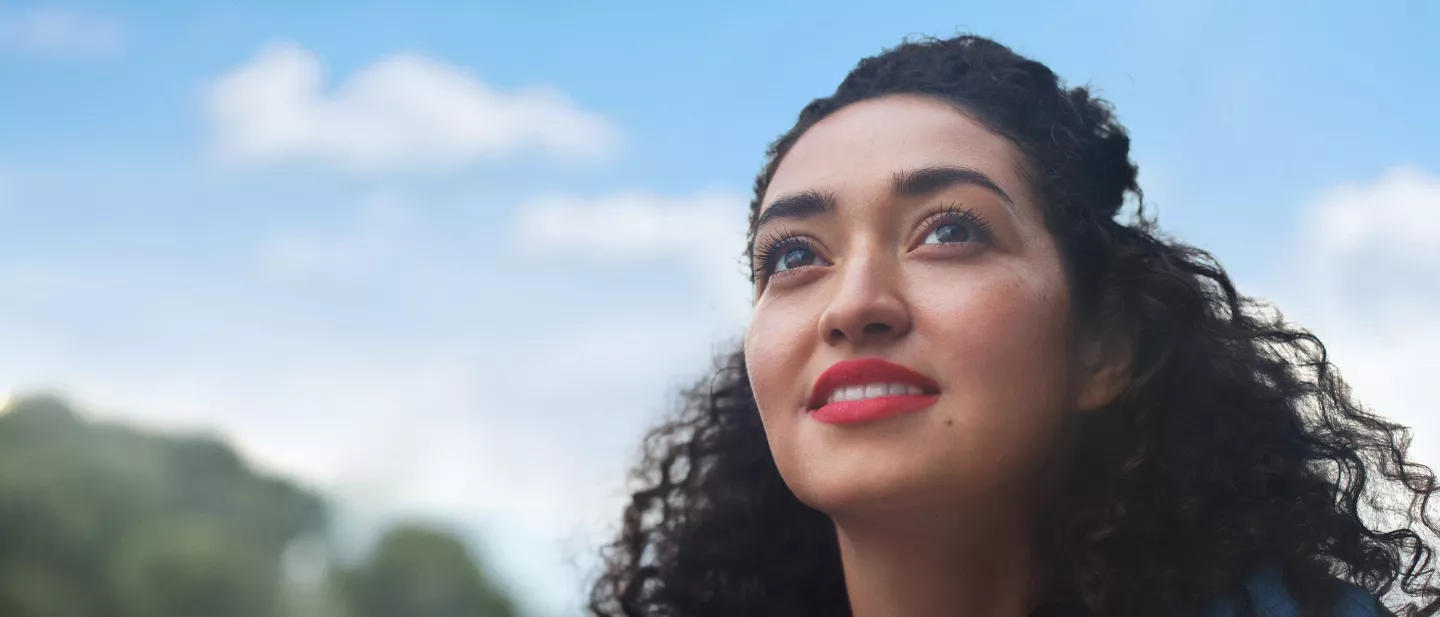 A woman with curly hair smiling and looking up outdoors against a blue sky