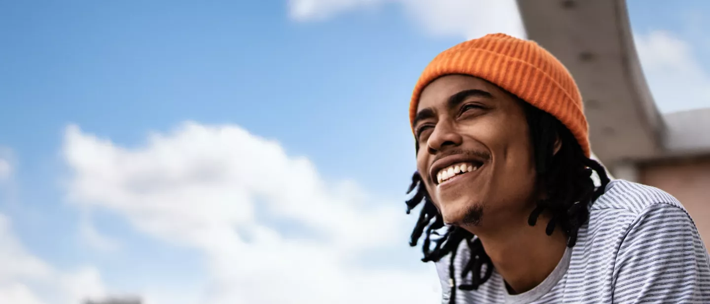 A young man with dreadlocks wearing an orange beanie, smiling on a rooftop