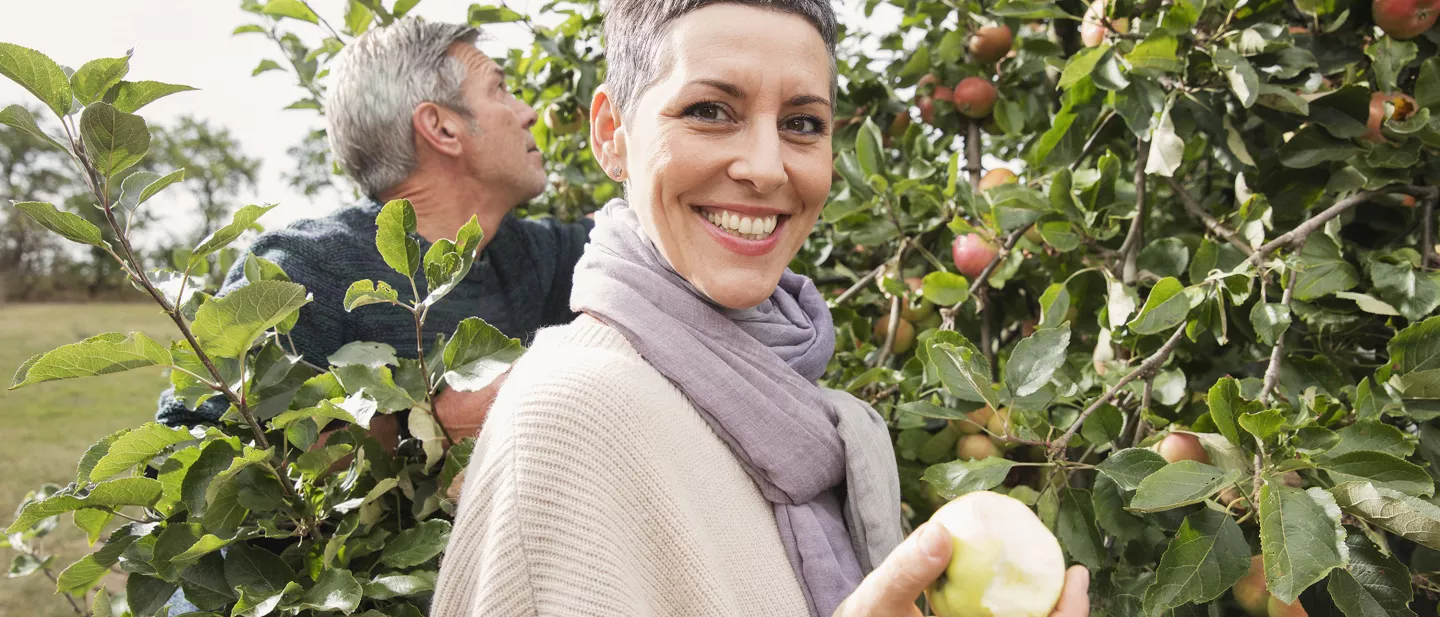 Smiling woman holding an apple in an orchard with another person picking apples behind her