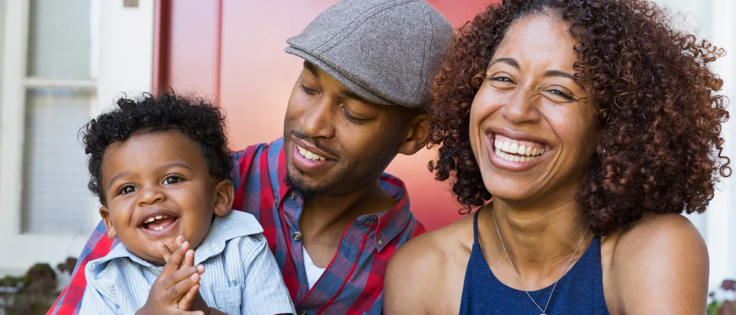 Smiling parents sit with their cheerful baby outside a red door.