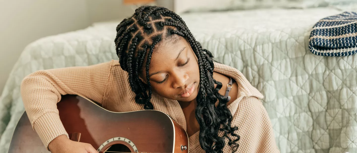 Girl playing acoustic guitar in a cozy bedroom setting.