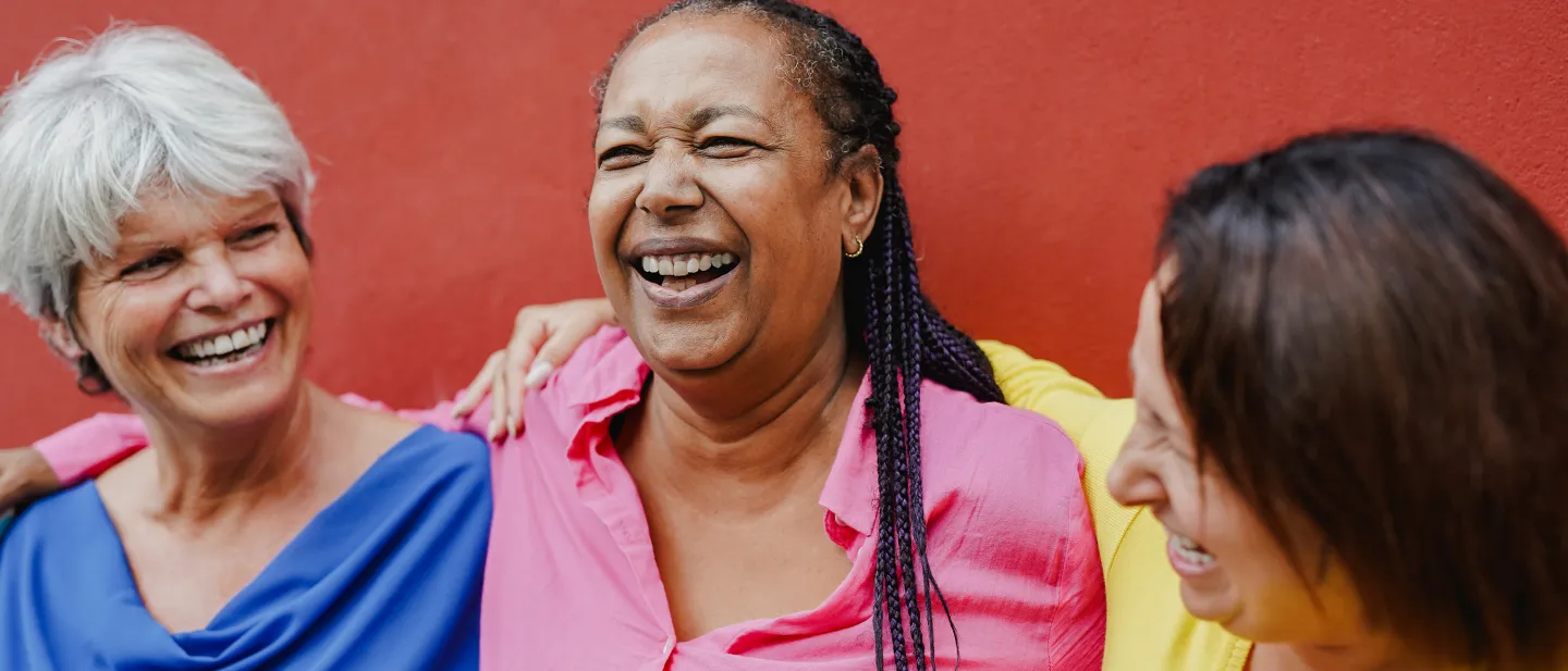 Three women laughing together with arms around each other, standing in front of a bright red wall.