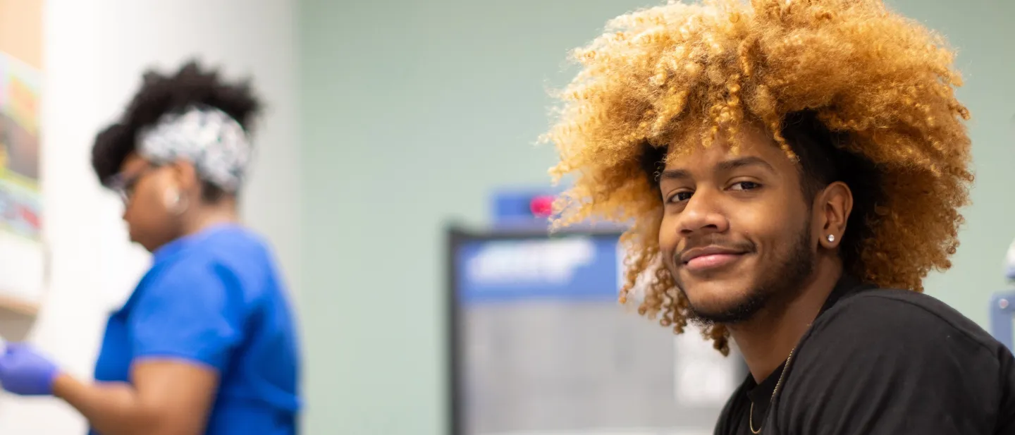 Young man with curly hair smiles while waiting in a medical setting, nurse working in background.