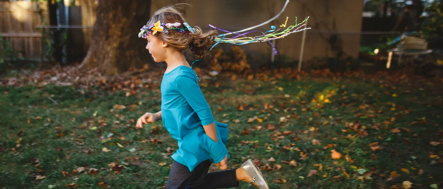 Young girl wearing a blue top and a colorful ribbon headband runs joyfully through a backyard filled with fallen leaves.