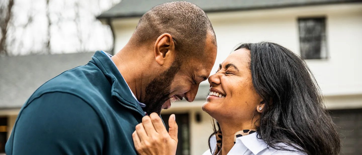 Smiling couple touching foreheads and laughing in front of a house, sharing a joyful and intimate moment.