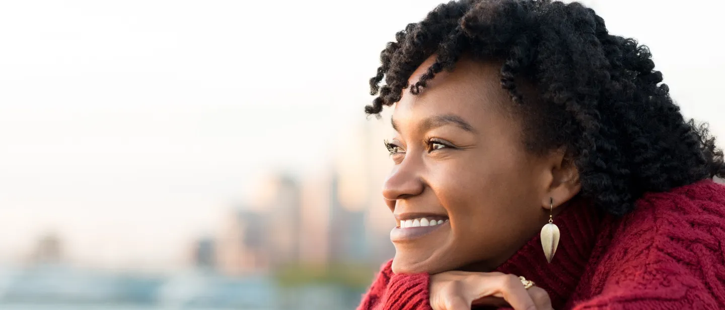 Smiling woman in red sweater looking out over water with city skyline in background.