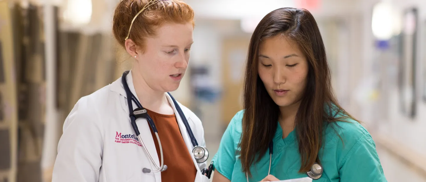 Two healthcare professionals in scrubs and a white coat discuss medical charts in a hospital hallway.