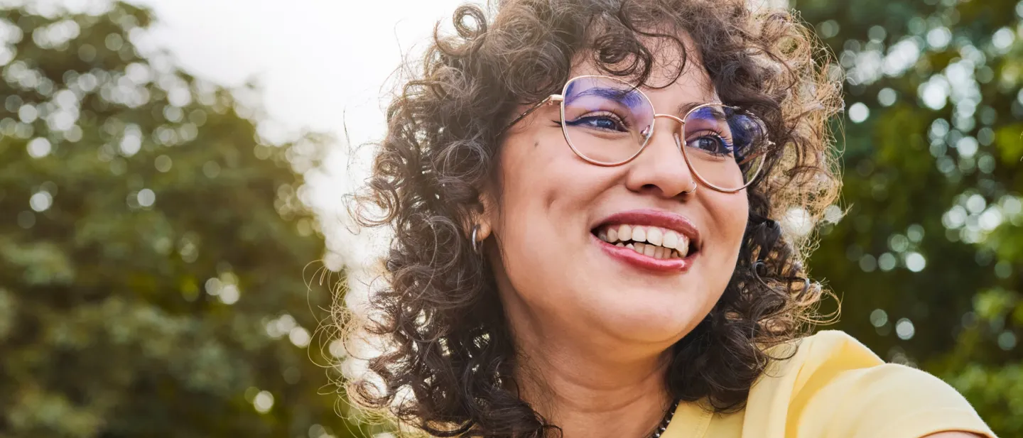 Woman wearing glasses with a big smile with trees in the background.