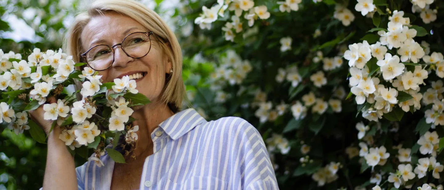 Woman in a garden enjoying being surrounded by small white flowers, smiling, wearing glasses