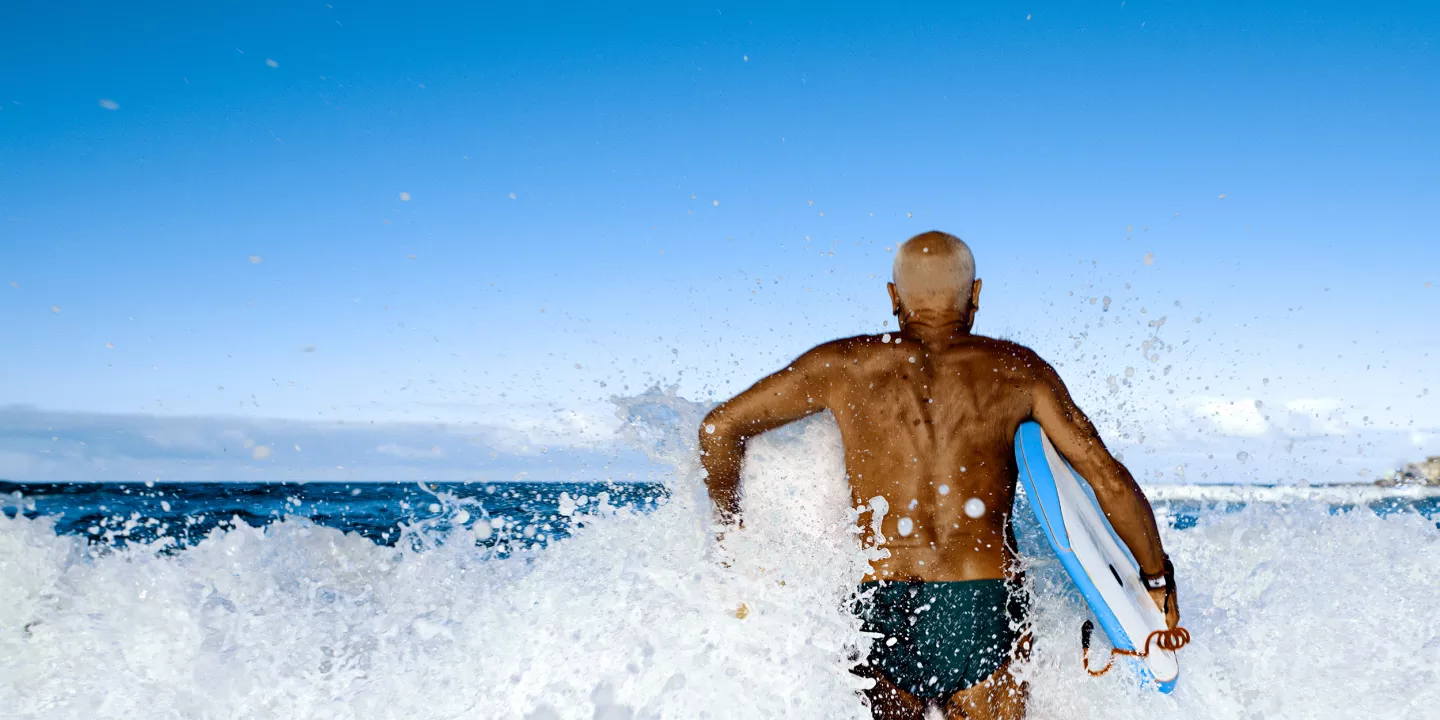 An older man walking in the waves with a bodyboard.