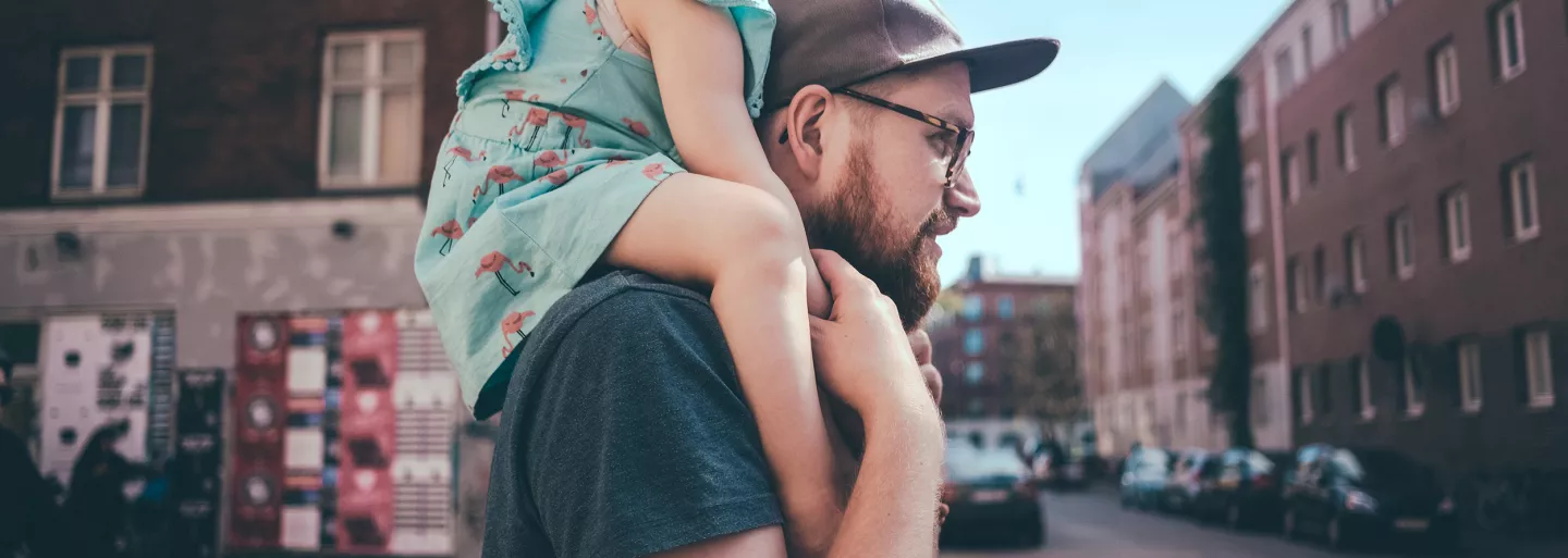 Side shot of a younger father carrying his daughter on his shoulders down a city street.