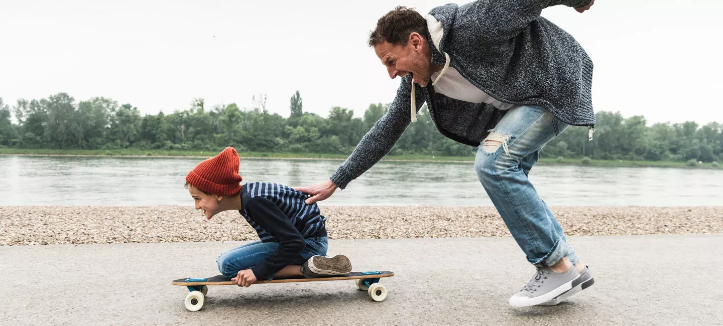 A happy father pushing son on skateboard by a river.