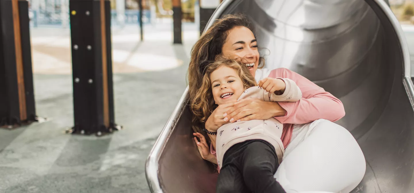 Young happy mother with her daughter having fun in tube slide on playground. 
