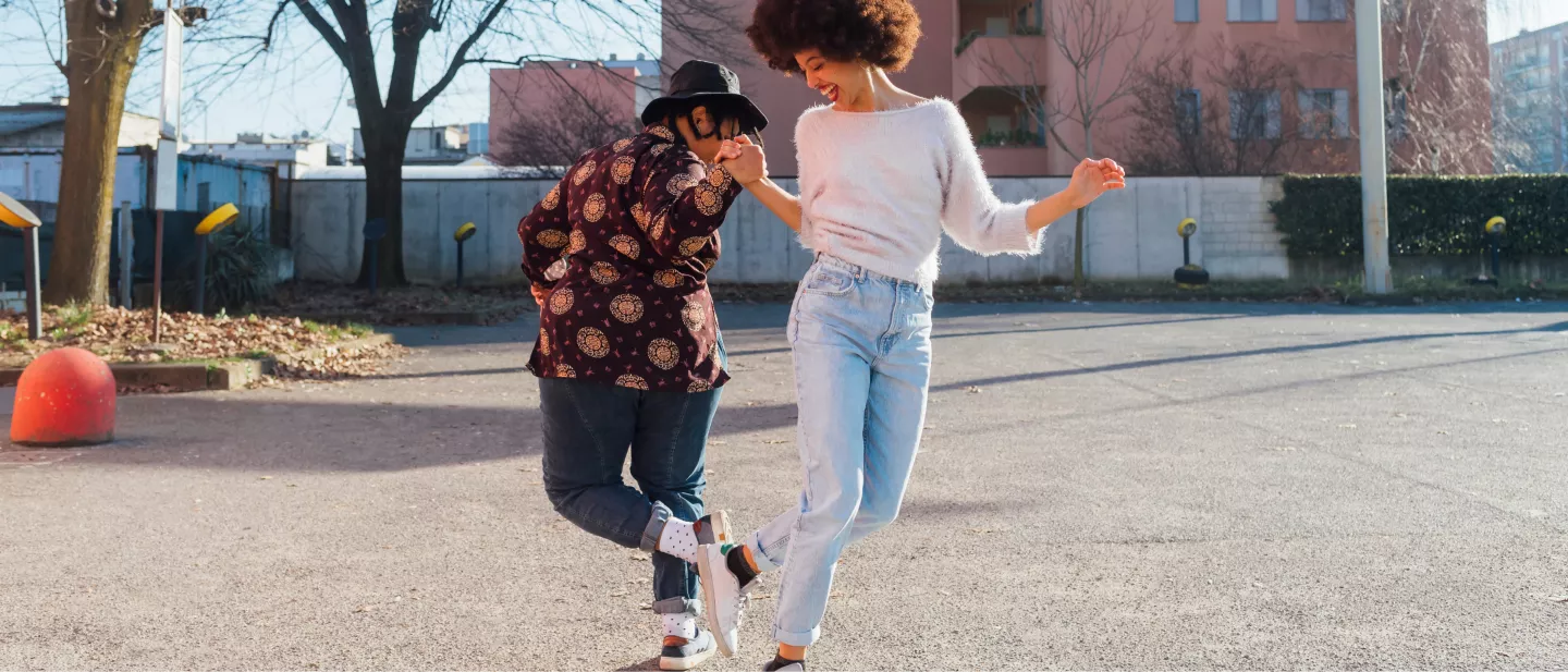 Two stylish friends dance in a parking lot near a green space full of trees.