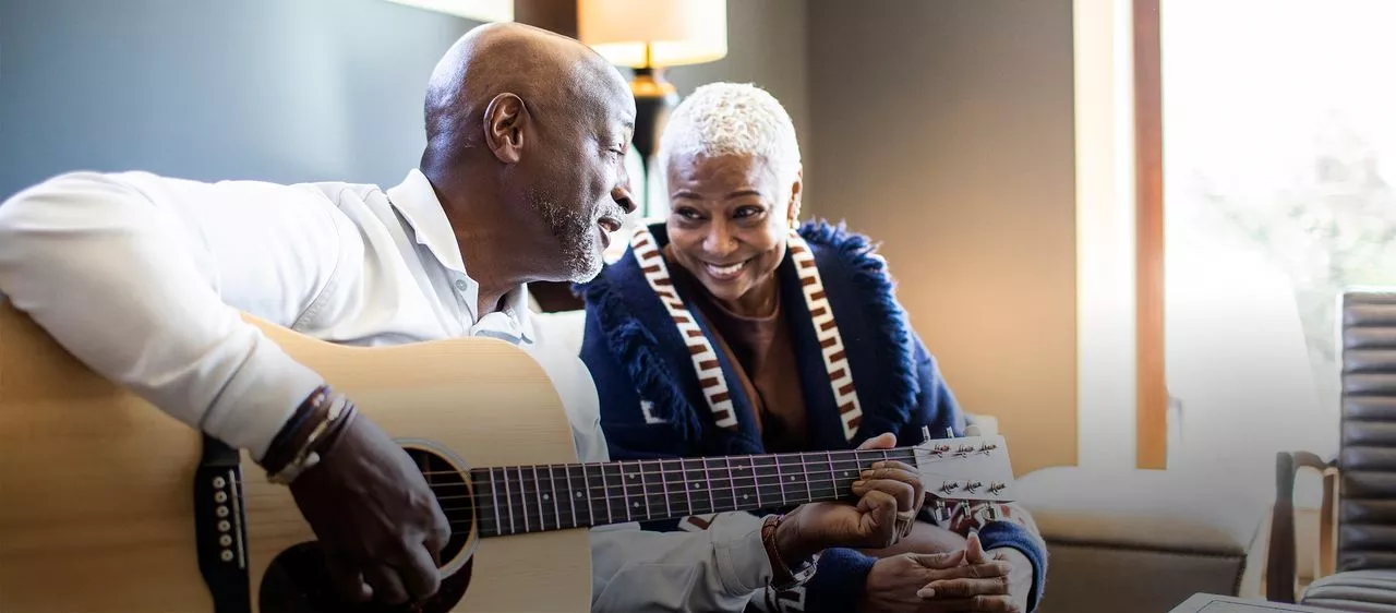 An older couple sit together in a comfortable room. One plays a guitar while the other smiles.