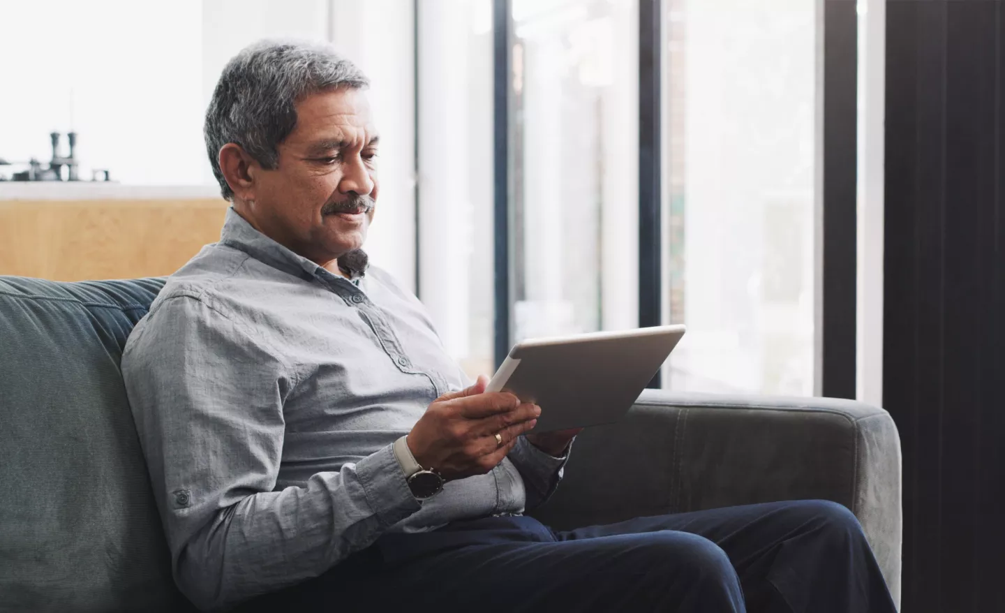 Man seated on couch holding tablet near window