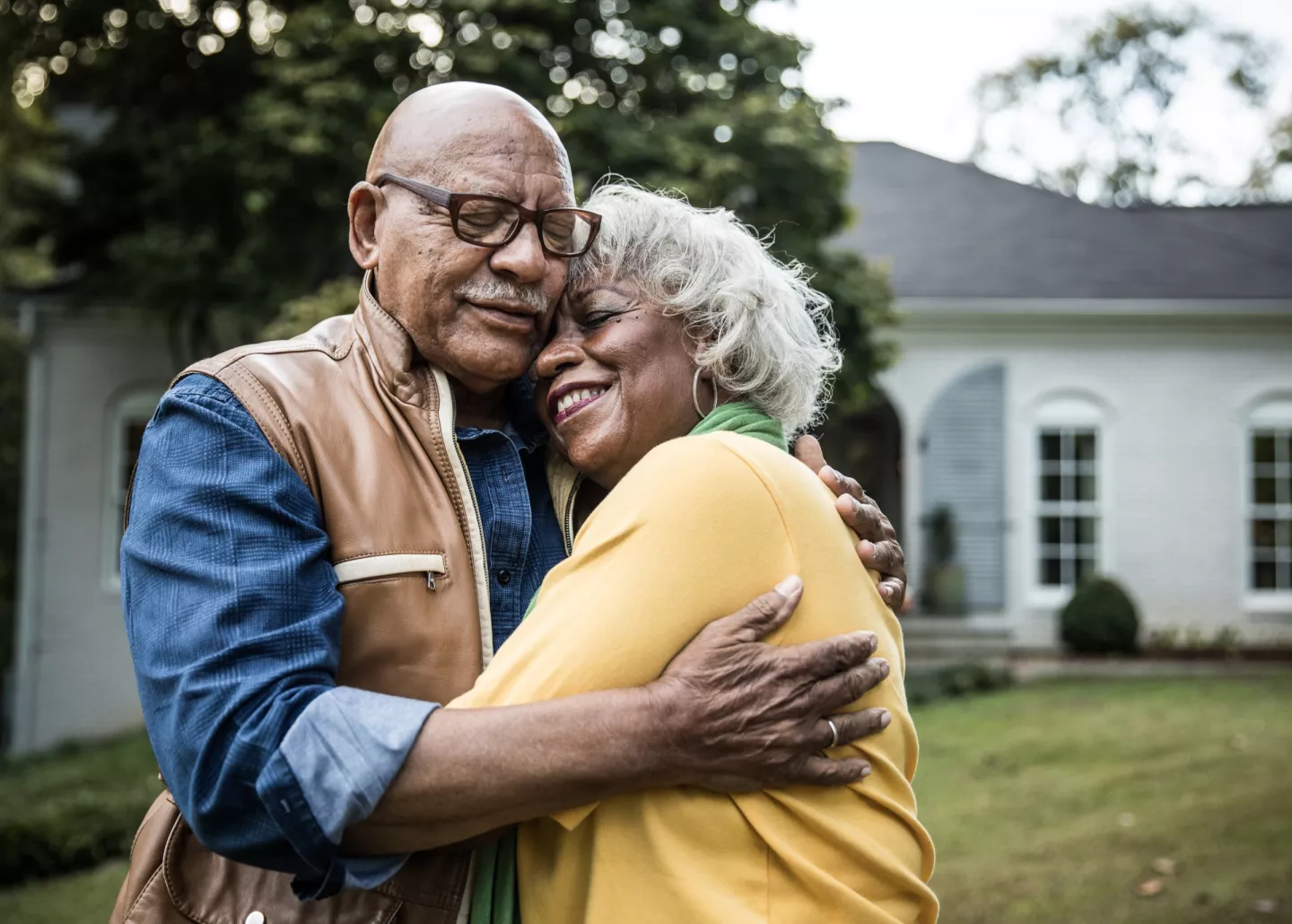 Senior couple smiling while embracing outdoors in front yard of home.