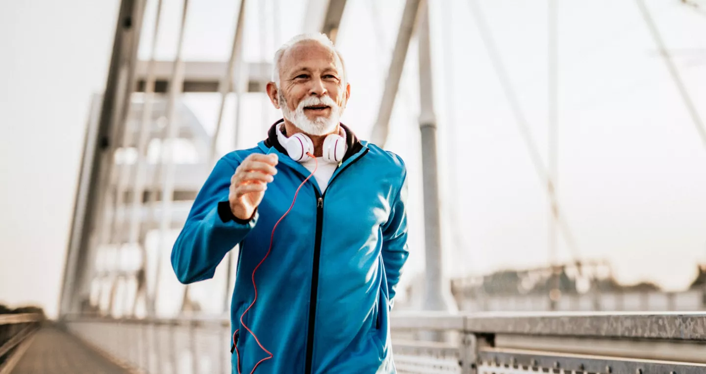 Senior gentleman in running outfit with headphones around neck jogging across bridge path