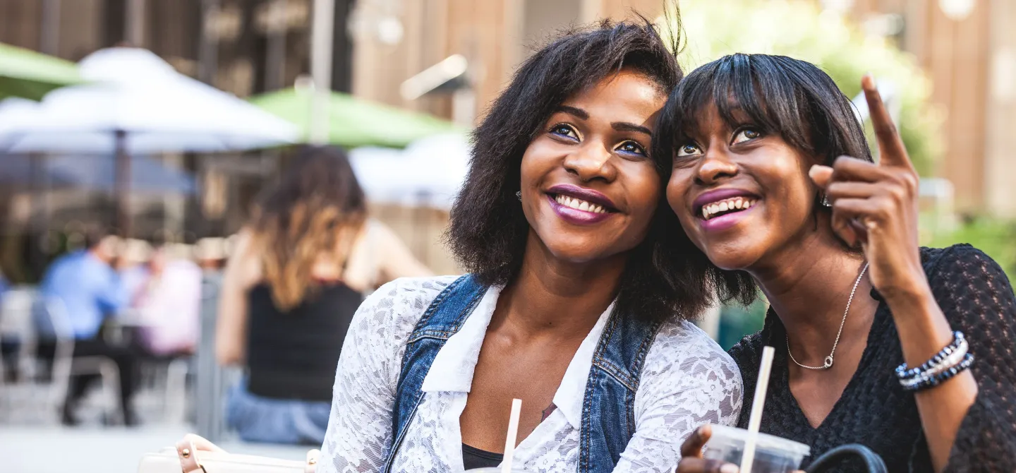 Two smiling women enjoying drinks outdoors while one points up excitedly.