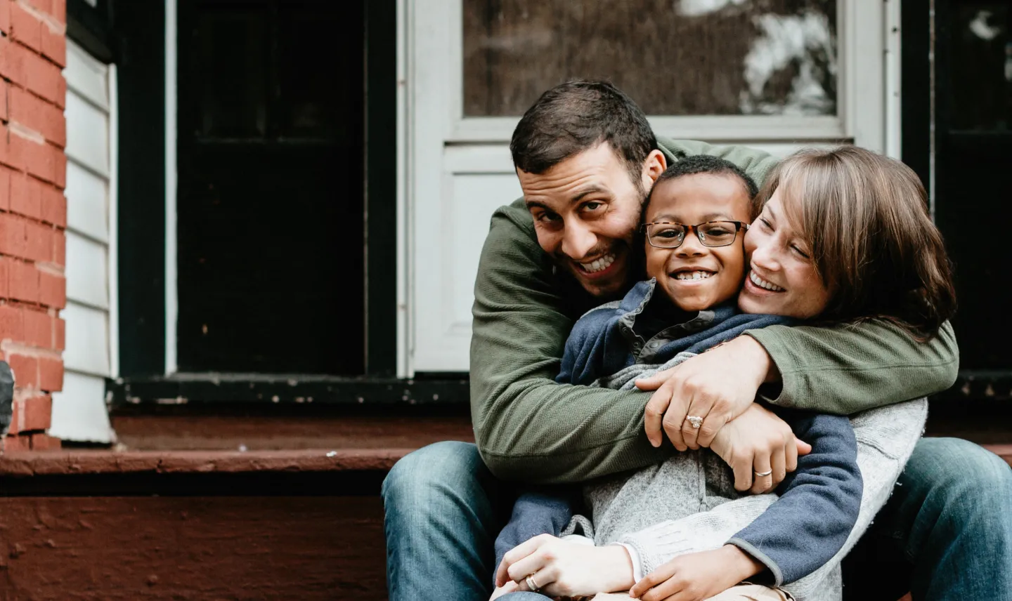 Smiling parents hugging their child while sitting on the front steps of their home.